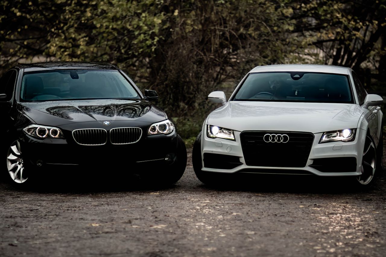 Black BMW and white Audi luxury sedans parked outdoors on a gravel path.